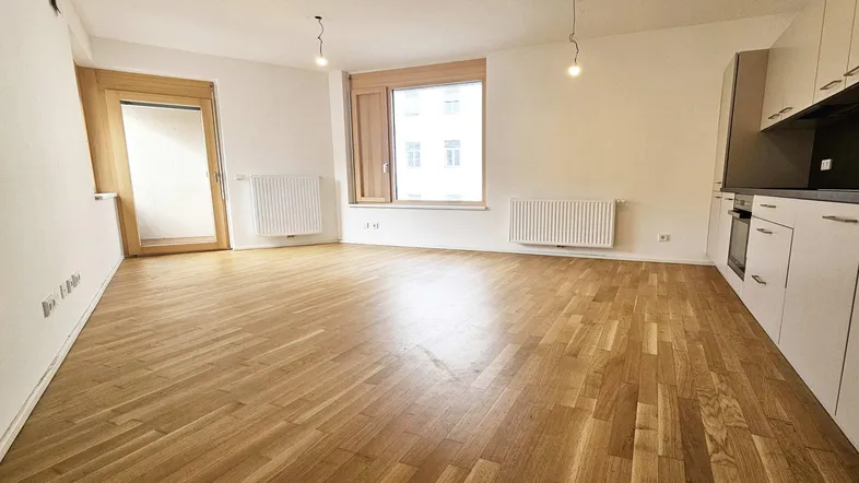 Heller offener Wohnbereich mit Parkettboden, großen Fenstern und Zugang zum Balkon.