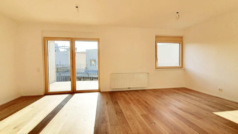 Helles, leeres Zimmer mit Holzboden, großem Fenster und Zugang zu einem Balkon, ideal als Wohnzimmer.