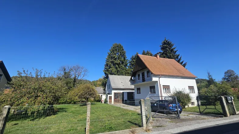 Weißes Einfamilienhaus mit rotem Dach, umzäuntem Vorgarten und Einfahrt unter blauem Himmel.