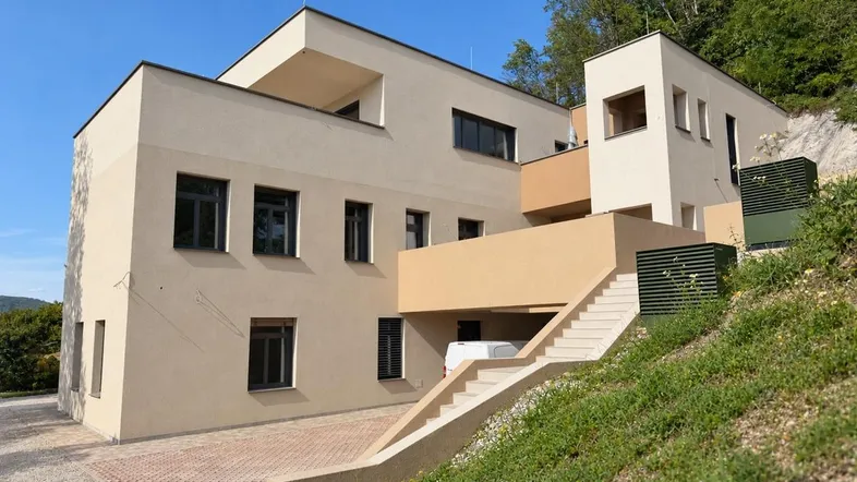 Moderne Architektur mit beigefarbener Fassade und großen Fenstern, eingebettet in eine grüne Hanglage.