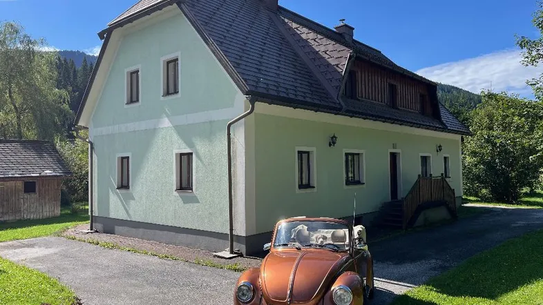 Traditionelles Landhaus mit hellgrüner Fassade und Satteldach, umgeben von Natur und einem Oldtimer auf der Auffahrt.