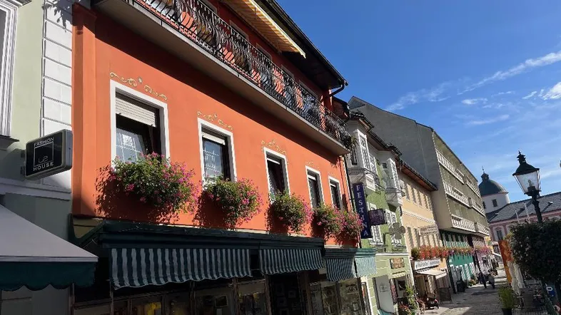 Lebhaftes orangefarbenes Gebäude mit Blumenkästen und Balkon in einer belebten Straße unter blauem Himmel.