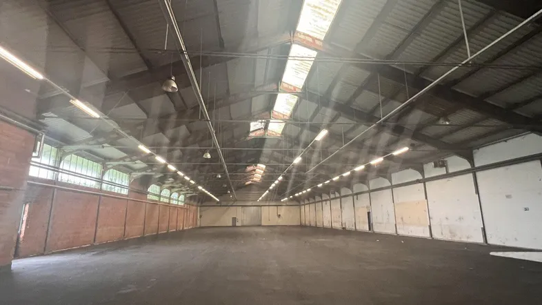 Große, leere Lagerhalle mit hohen Decken, Oberlichtern und Ziegelwänden.