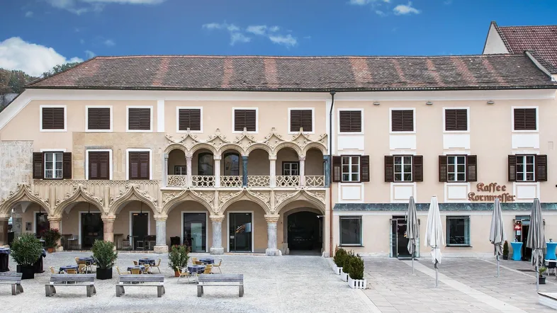 Historisches Gebäude mit aufwendiger Fassade, Arkaden und einem Café im Erdgeschoss an einem öffentlichen Platz.