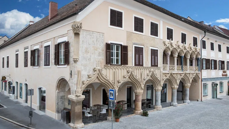 Historisches Gebäude mit aufwendiger Fassade, Arkaden und Balkonen an einem gepflasterten Platz.