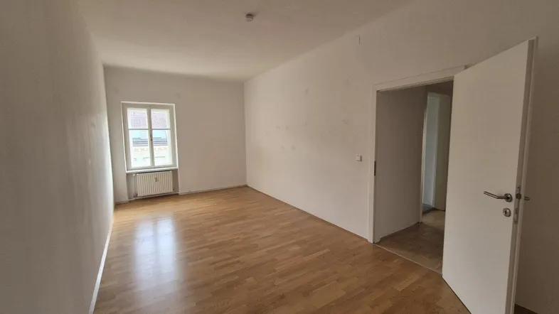 Helles, leeres Zimmer mit Parkettboden und einem Fenster, das Tageslicht hereinlässt.