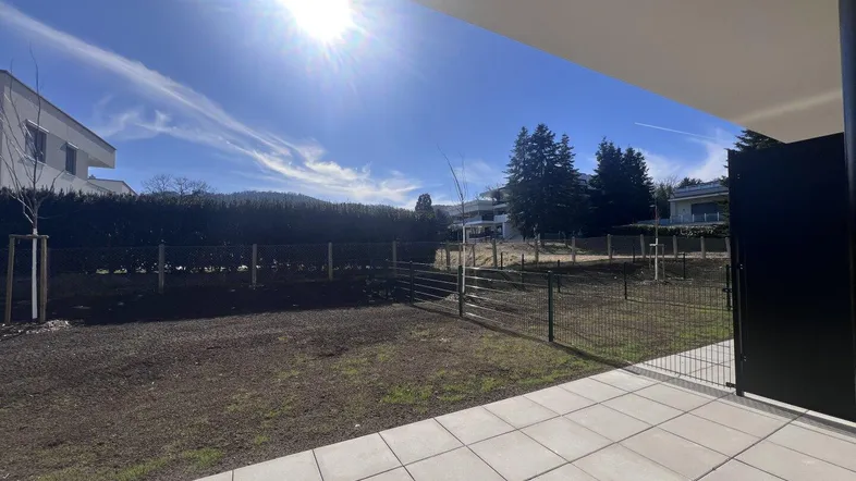 Sonnige Terrasse mit Fliesenboden und angrenzendem Gartenbereich unter blauem Himmel.
