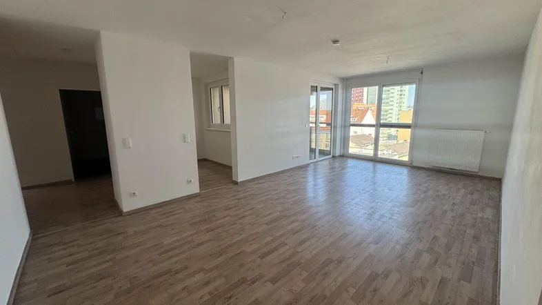 Geräumiges, leeres Wohnzimmer mit Laminatboden und großen Fenstern, die viel Tageslicht hereinlassen.
