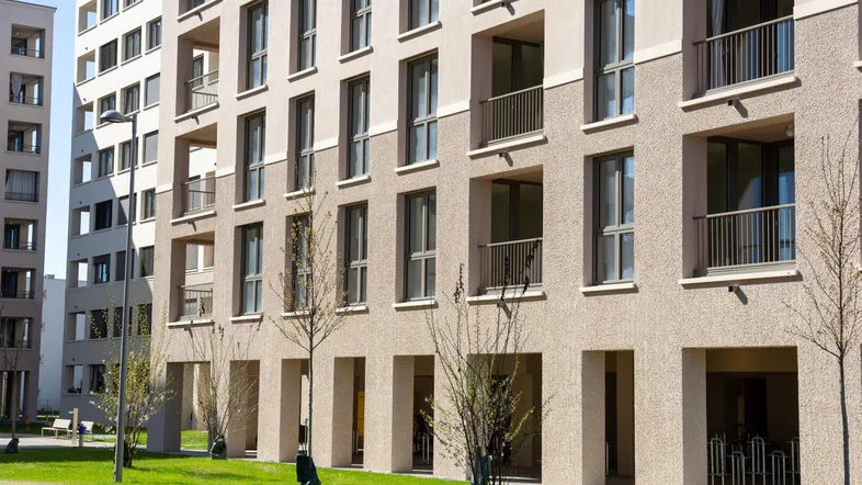 Moderne Wohngebäude-Fassade mit Balkonen, großen Fenstern und begrüntem Außenbereich im Sonnenlicht.