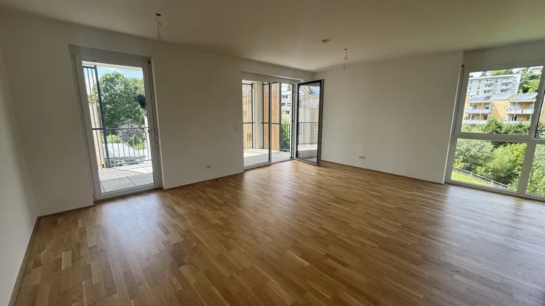 Helles Wohnzimmer mit Parkettboden, weißen Wänden und Zugang zu einem Balkon mit Grünblick.
