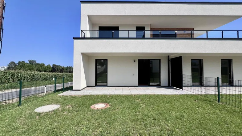 Modernes Neubaugebäude mit großer Terrasse und Gartenbereich, umgeben von grüner Landschaft.