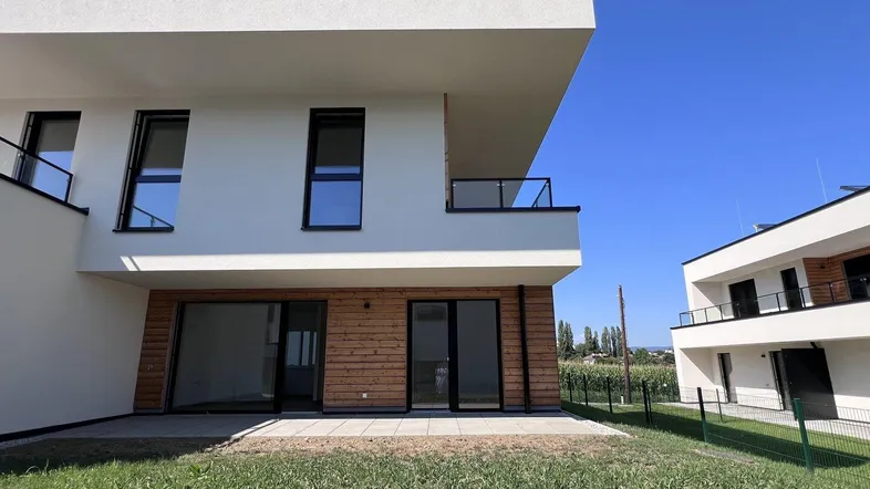 Moderne Neubauwohnung mit Terrasse und Gartenanteil, umgeben von grüner Landschaft unter blauem Himmel.