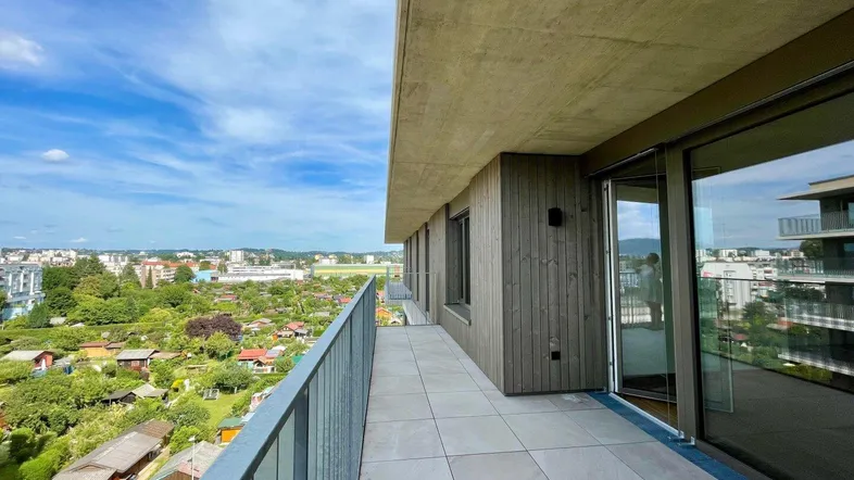 Großer Balkon mit Fliesenboden und weitem Blick über die Stadt und Schrebergärten unter blauem Himmel.
