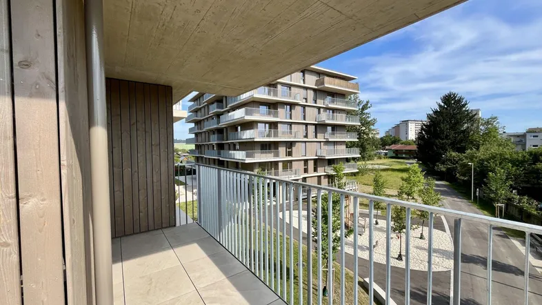 Blick von einem modernen Balkon mit Metallgeländer auf ein weiteres Gebäude und grüne Umgebung.