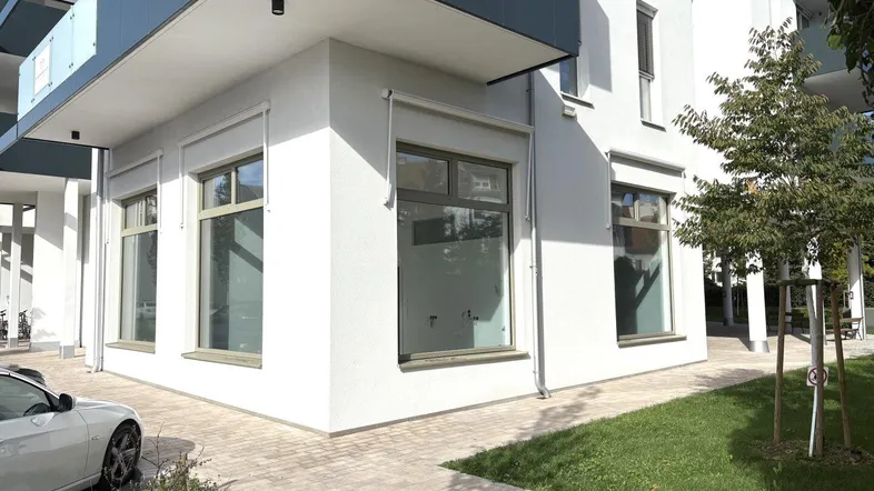 Moderne Geschäftsfassade im Erdgeschoss eines Neubaus mit großen Fenstern und gepflastertem Gehweg.