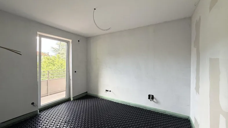 Unfertiger Raum mit sichtbarer Fußbodenheizung und Zugang zu einem Balkon, bereit für den Ausbau.