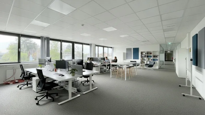 Großes, offenes Büro mit mehreren Arbeitsplätzen, Teppichboden und großen Fenstern für viel Tageslicht.