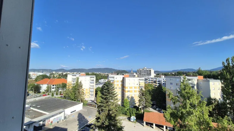 Weitblick über Wohngebäude und grüne Umgebung mit Bergen am Horizont unter blauem Himmel.