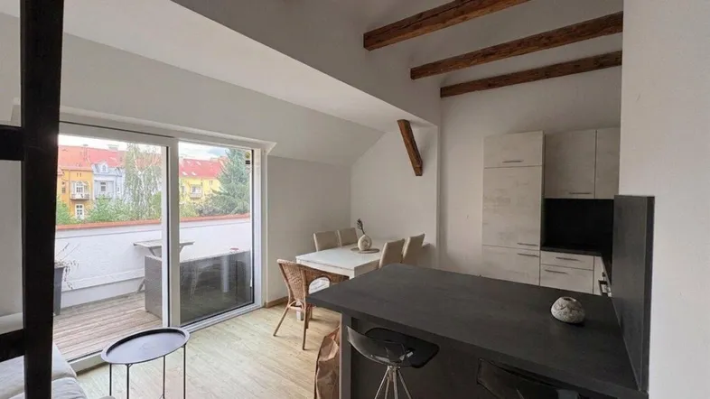 Heller offener Wohn- und Essbereich mit Holzbalken, Kücheninsel und Zugang zu einem Balkon.