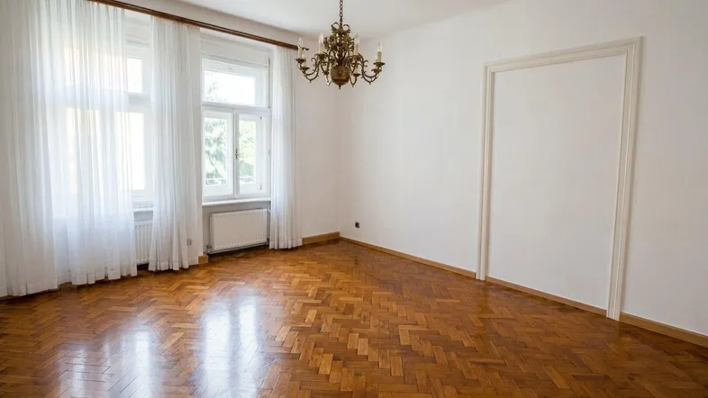 Helles Zimmer mit Fischgrätparkettboden, hohen Fenstern und einem klassischen Kronleuchter.