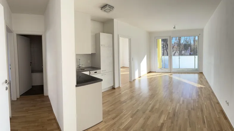 Helles, offenes Wohnzimmer mit integrierter Küche, Holzboden und Zugang zu einem Balkon.