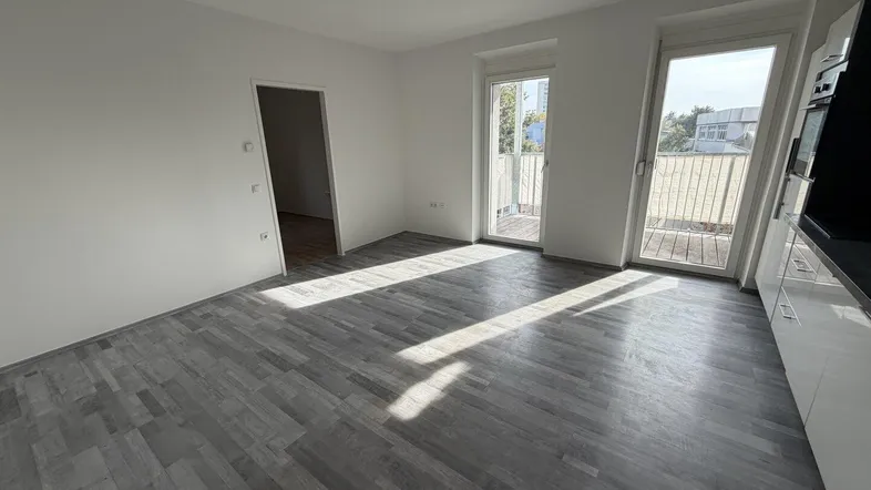 Helles Zimmer mit grauem Laminatboden und Zugang zu einem Balkon, durchflutet von Sonnenlicht.