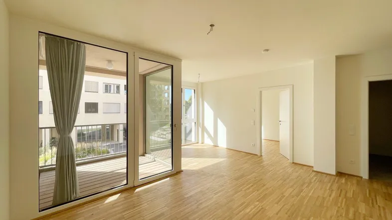 Heller, leerer Wohnbereich mit Holzboden und Zugang zu einem Balkon, durchflutet von Sonnenlicht.