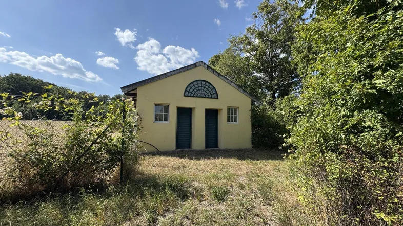 Kleines gelbes Gebäude mit grünen Türen und Rundbogenfenster, umgeben von Natur und Feldern.