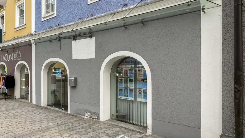 Graue Geschäftsfassade mit zwei großen Rundbogenfenstern und einem Glasvordach über dem Eingangsbereich zur Straße.