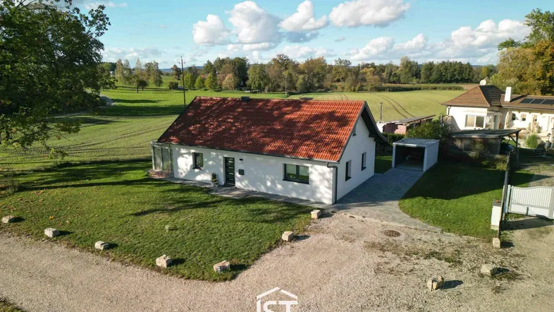 Freistehendes Einfamilienhaus mit rotem Ziegeldach, umgeben von grünen Feldern unter blauem Himmel.