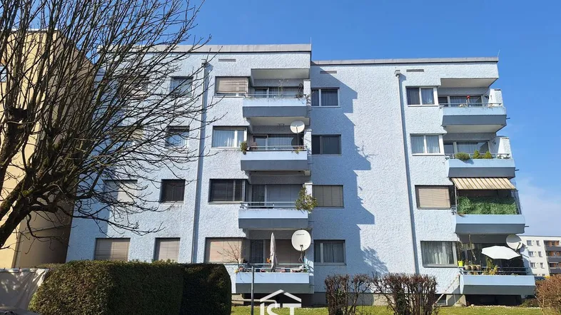Mehrfamilienhaus mit hellblauer Fassade, mehreren Balkonen und gepflegter Grünanlage unter blauem Himmel.