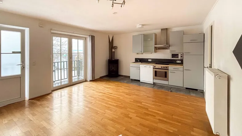 Offener Wohnbereich mit integrierter Küche, Holzboden und Zugang zu einem Balkon. Ein Kaminofen sorgt für Gemütlichkeit.
