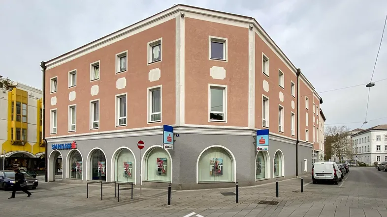 Eckgebäude mit traditioneller Architektur, rosa Fassade und Geschäftsräumen im Erdgeschoss an einer Stadtstraße.