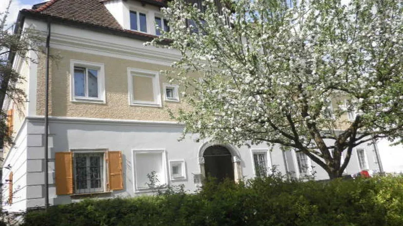 Historisches Gebäude mit blühendem Baum im Vordergrund und einem gewölbten Eingangsbereich.