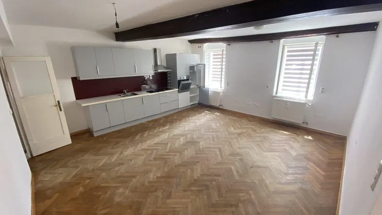 Geräumiger Wohnbereich mit integrierter Küche, sichtbaren Holzbalken und Fischgrätparkettboden.
