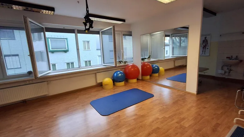Geräumiger Therapieraum mit Holzboden, großen Fenstern und Sportgeräten für Übungen und Behandlungen.