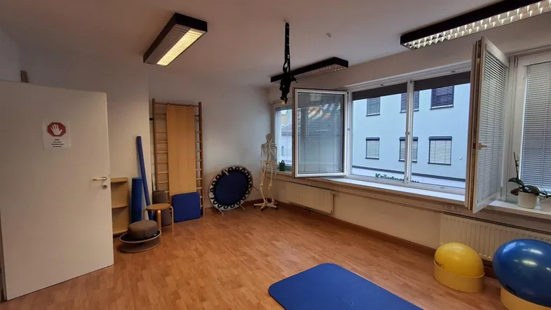 Heller Therapieraum mit Holzboden, Sprossenwand und medizinischen Modellen für Behandlungen und Training.