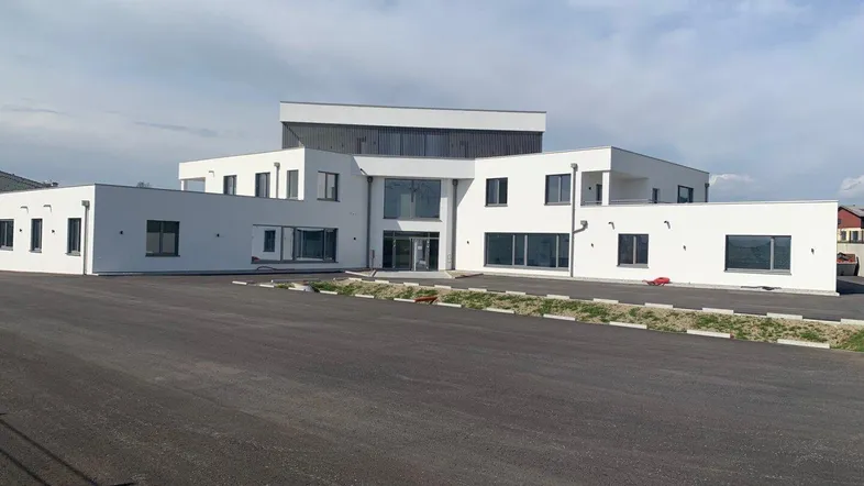 Modernes, weißes Gebäude mit großen Fenstern und einem weitläufigen Parkplatz davor unter bewölktem Himmel.