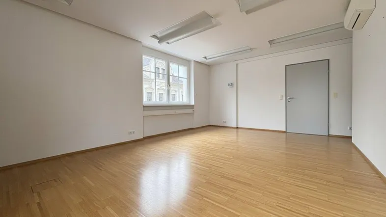Geräumiges Büro mit hellem Parkettboden, weißen Wänden und einem Fenster mit Blick nach draußen.