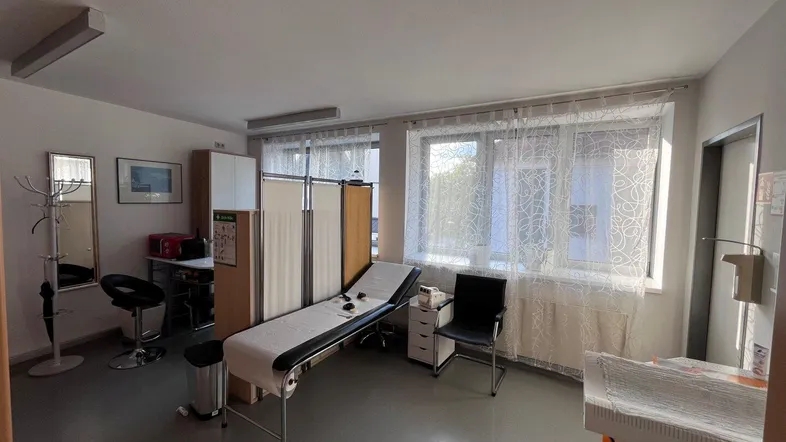 Heller Untersuchungsraum mit Liege, Fensterfront und Sichtschutzwänden, ideal für eine Arztpraxis.