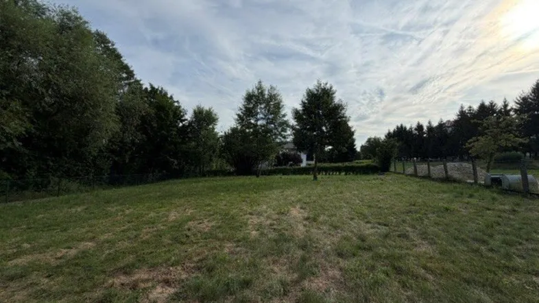 Weitläufige Grünfläche mit Bäumen im Hintergrund und einem Haus in der Ferne unter bewölktem Himmel.