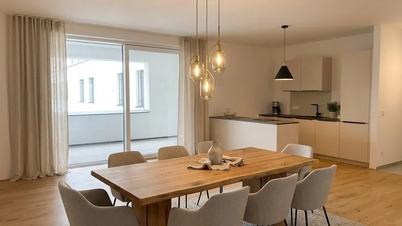Heller offener Wohn-Essbereich mit Holztisch, modernen Pendelleuchten und Zugang zu einem Balkon.