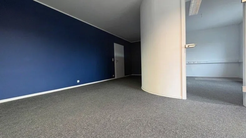 Geräumiges Büro mit dunkelblauer Akzentwand, grauem Teppichboden und Glastür zu einem weiteren Raum.