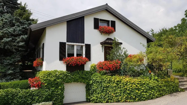 Weißes Einfamilienhaus mit dunklem Dach, grüner Bepflanzung und roten Blumenkästen.