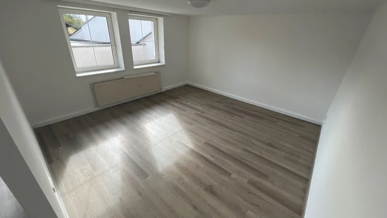 Helles Zimmer mit modernem Laminatboden, weißen Wänden und zwei Fenstern, die Tageslicht hereinlassen.