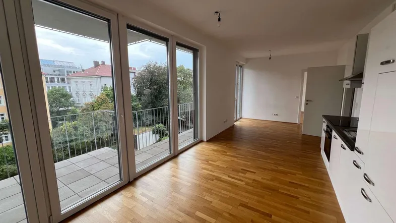 Heller offener Wohnbereich mit Holzboden, integrierter Küche und Zugang zu einem Balkon mit Blick ins Grüne.