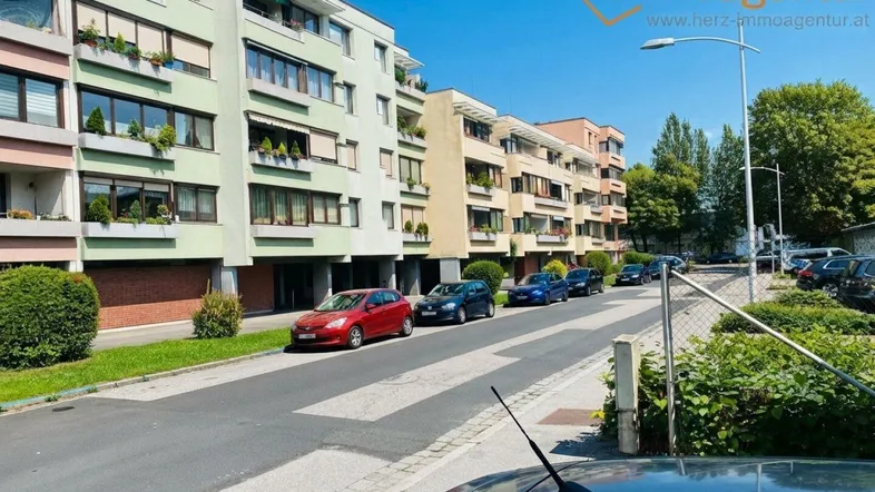 Mehrere moderne Wohngebäude entlang einer Straße mit geparkten Autos und gepflegten Grünflächen.