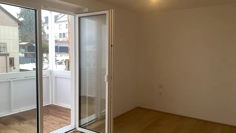Helles Zimmer mit Holzboden und großen Glasfenstern, die Zugang zu einem kleinen Balkon bieten.