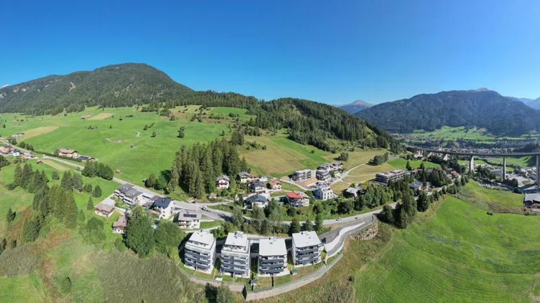 Panoramablick auf das Tal mit modernen Gebäuden, umgeben von grünen Wiesen und majestätischen Bergen.