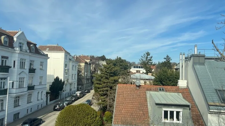 Blick über eine ruhige Wohnstraße mit geparkten Autos und traditionellen Gebäuden unter blauem Himmel.